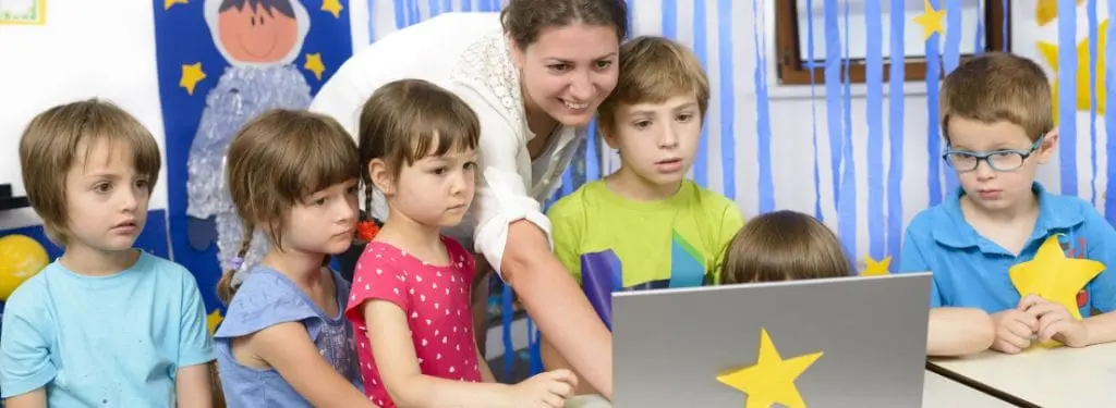 a daycare teacher surrounded by children while checking a presentation on a laptop using a daycare software|Brightwheel|HiMama|iCare|Jackrabbit Care|Kangarootime|LifeCubby|Procare Solutions|Sandbox|Sawyer