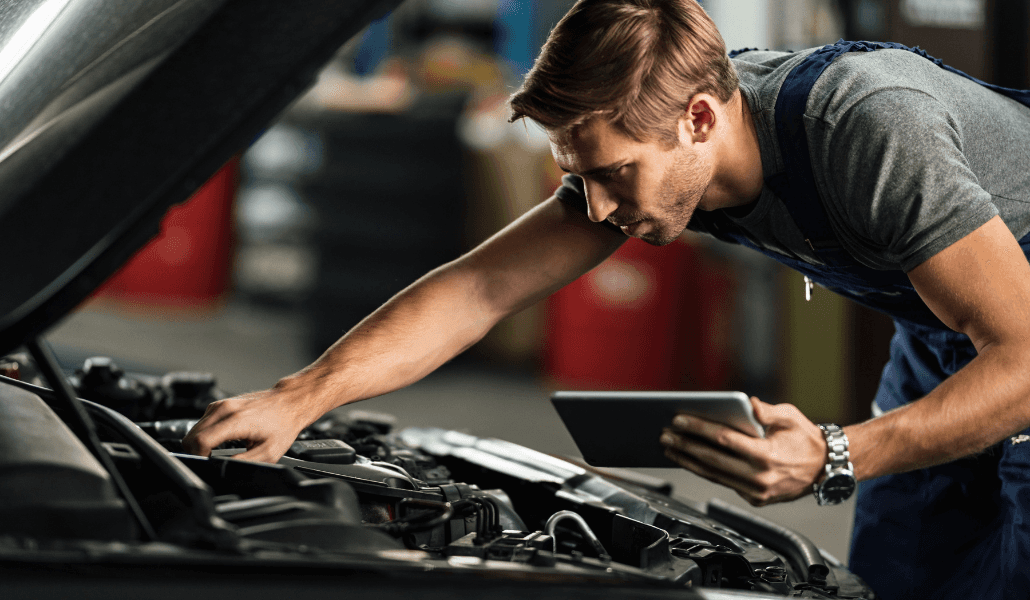 A mechanic inspects a car engine while holding a tablet in an auto repair shop.