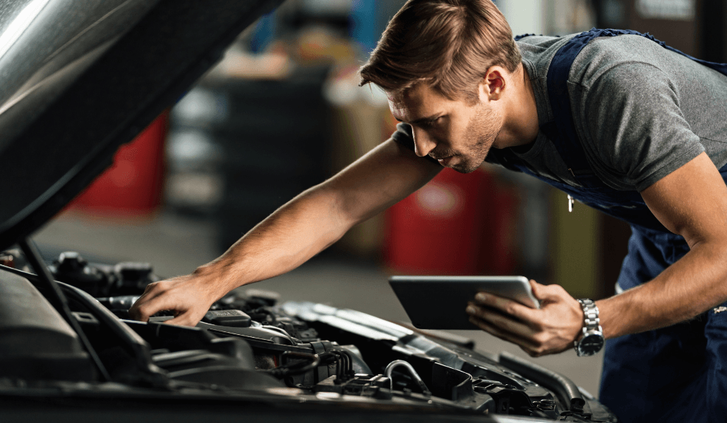 A mechanic inspects a car engine while holding a tablet in an auto repair shop.