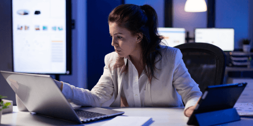 A woman in a white blazer works at a laptop late at night in a dimly lit office, with a presentation screen visible in the background.