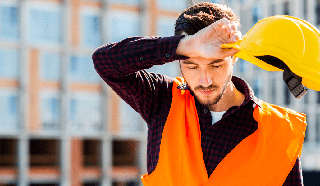 A tired construction worker in an orange safety vest wipes his forehead from sweat while holding a yellow hard hat at a building site.