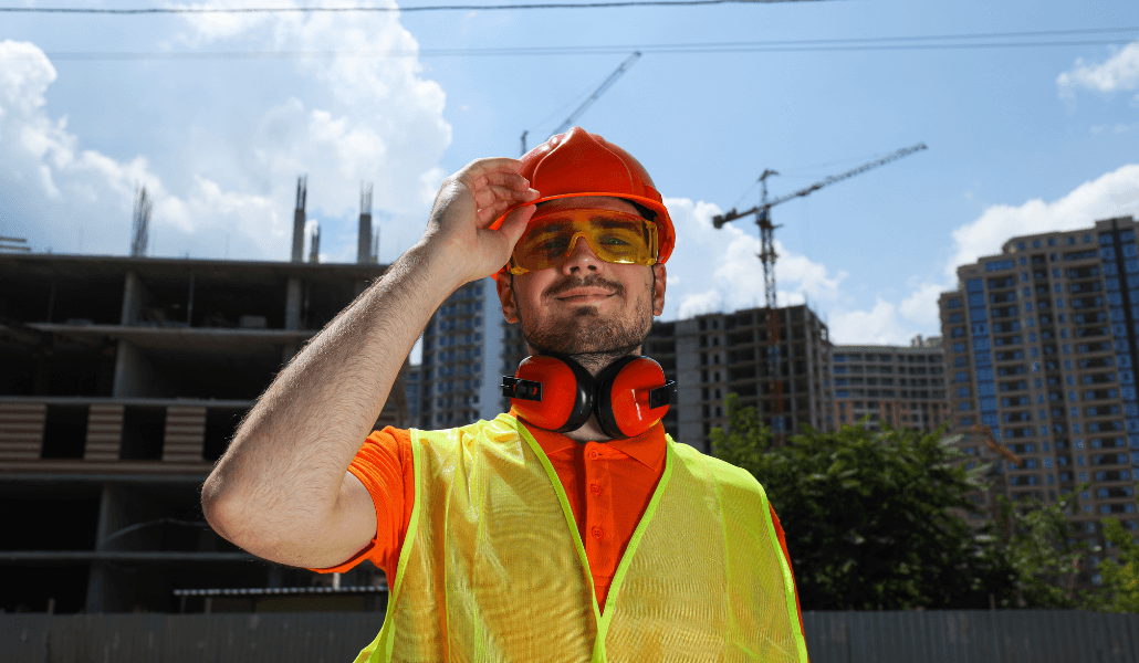 Construction worker in orange safety gear shielding his eyes from the sun on a job site.