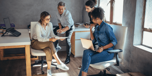 Team of four colleagues collaborating in an office, reviewing documents and using laptops.