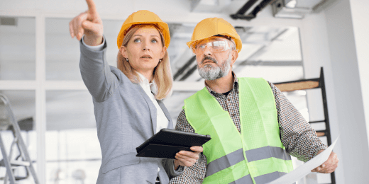 Two construction professionals wearing hard hats and safety gear reviewing building plans and pointing at something on site.