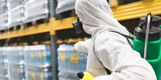 An exterminator in a full-body coverall and gas mask relies on standard PPE for pest control to safely spray a shelving area.