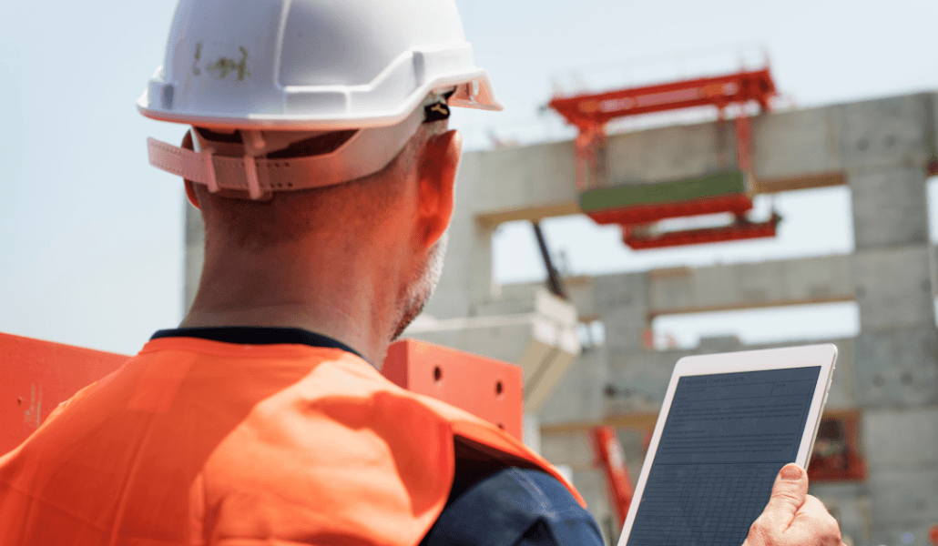 A construction worker in a hard hat and high-vis vest using a tablet on an active building site.