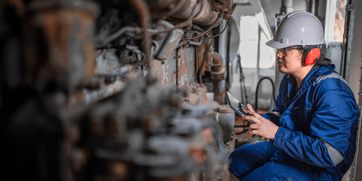 Engineer wearing protective gear inspects industrial machinery while using a tablet.