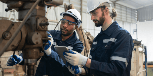 Two factory workers in safety gear inspecting machinery together with a tablet.