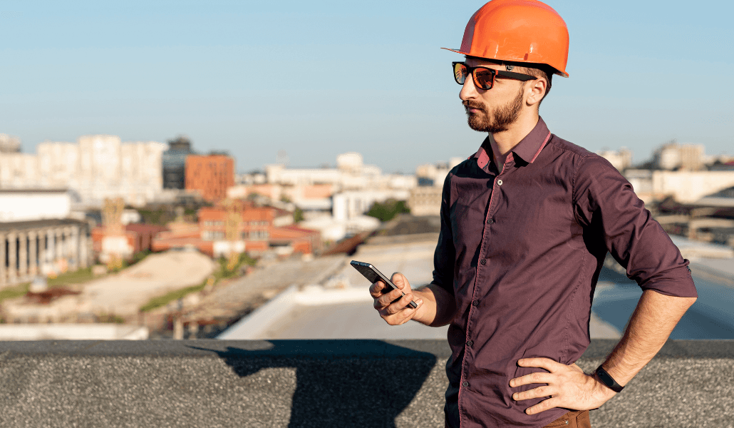 A site manager in a hard hat and sunglasses checking his phone on a rooftop worksite.