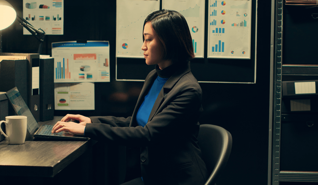 Woman in a blazer reviewing risk data on a laptop, surrounded by charts and dashboards on the wall behind her.