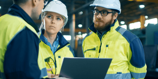 Three engineers in safety gear discussing team tasks while looking at a laptop on a factory floor.