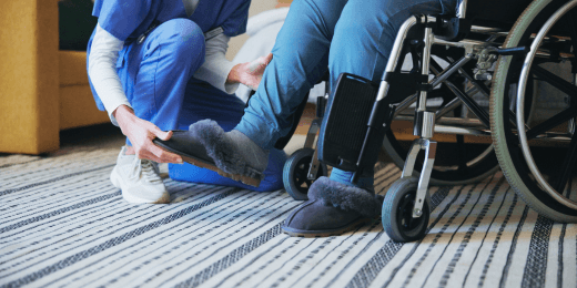 Care worker assisting a person in a wheelchair with their slippers indoors.