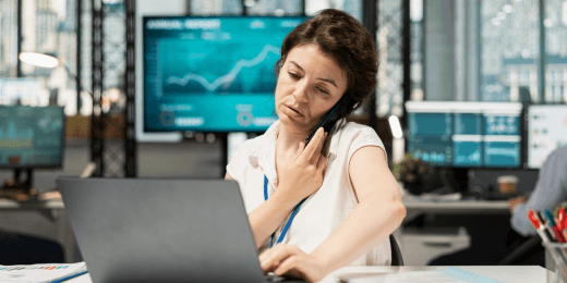 Businesswoman on a phone call while reviewing data on a laptop, with financial charts displayed on a screen behind her.
