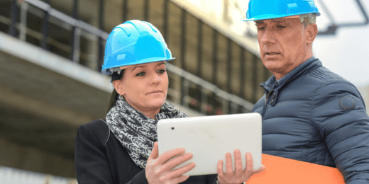 Two construction professionals wearing blue hard hats review information on a tablet at a worksite.