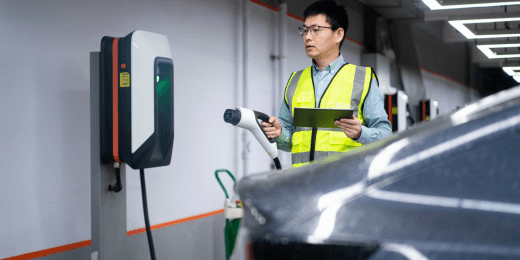 Engineer holding a charging cable and tablet beside an electric vehicle charging station in a parking garage.