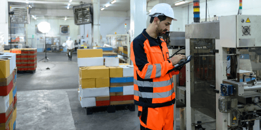 Worker in high-visibility safety gear and hard hat inspecting machinery in a factory using a tablet.
