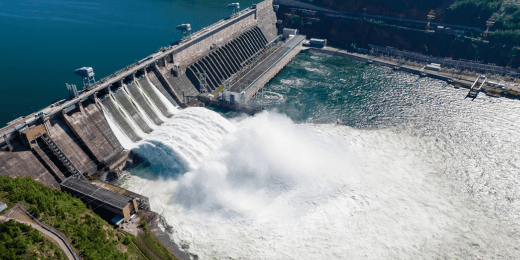 An aerial view of a massive concrete dam generating hydroelectric power as water surges through open spillways into the river below.