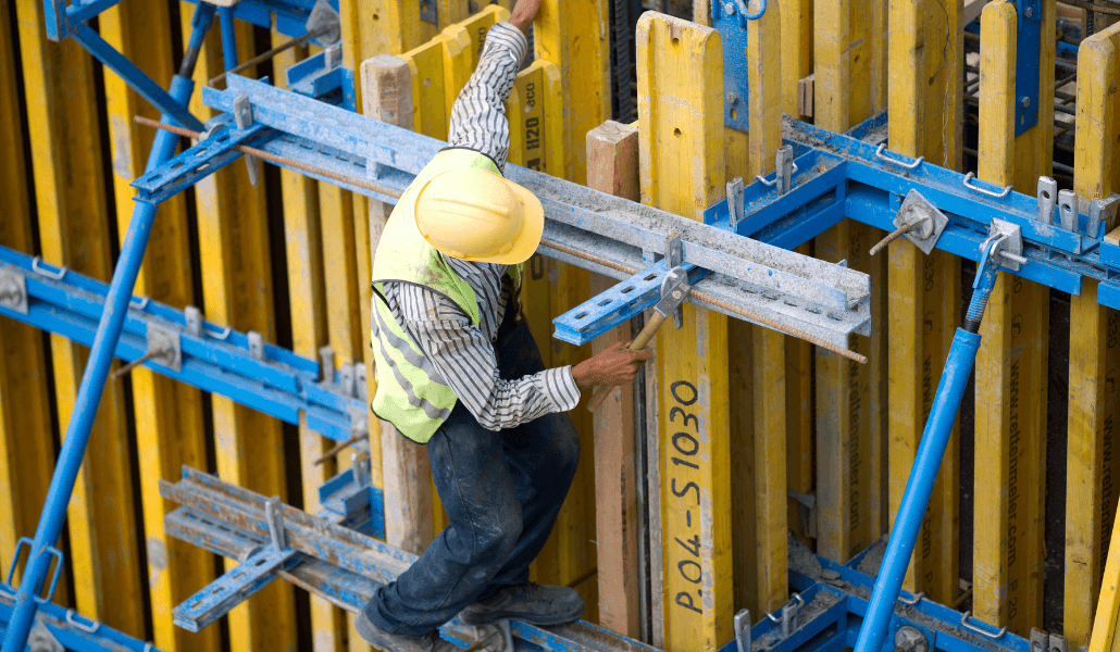 Construction worker in a yellow hard hat and safety vest working on scaffolding at a building site.