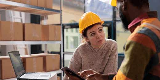 Two warehouse workers reviewing supplier risks, one holding a tablet near a laptop with shelves of boxes behind them.