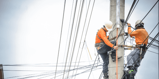 Two utility workers wearing safety harnesses and orange shirts climb a power pole to repair electrical wires.