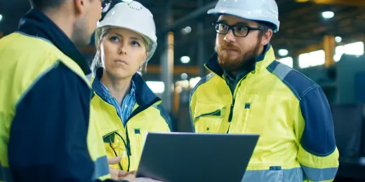 Three engineers in safety gear discussing team tasks while looking at a laptop on a factory floor.